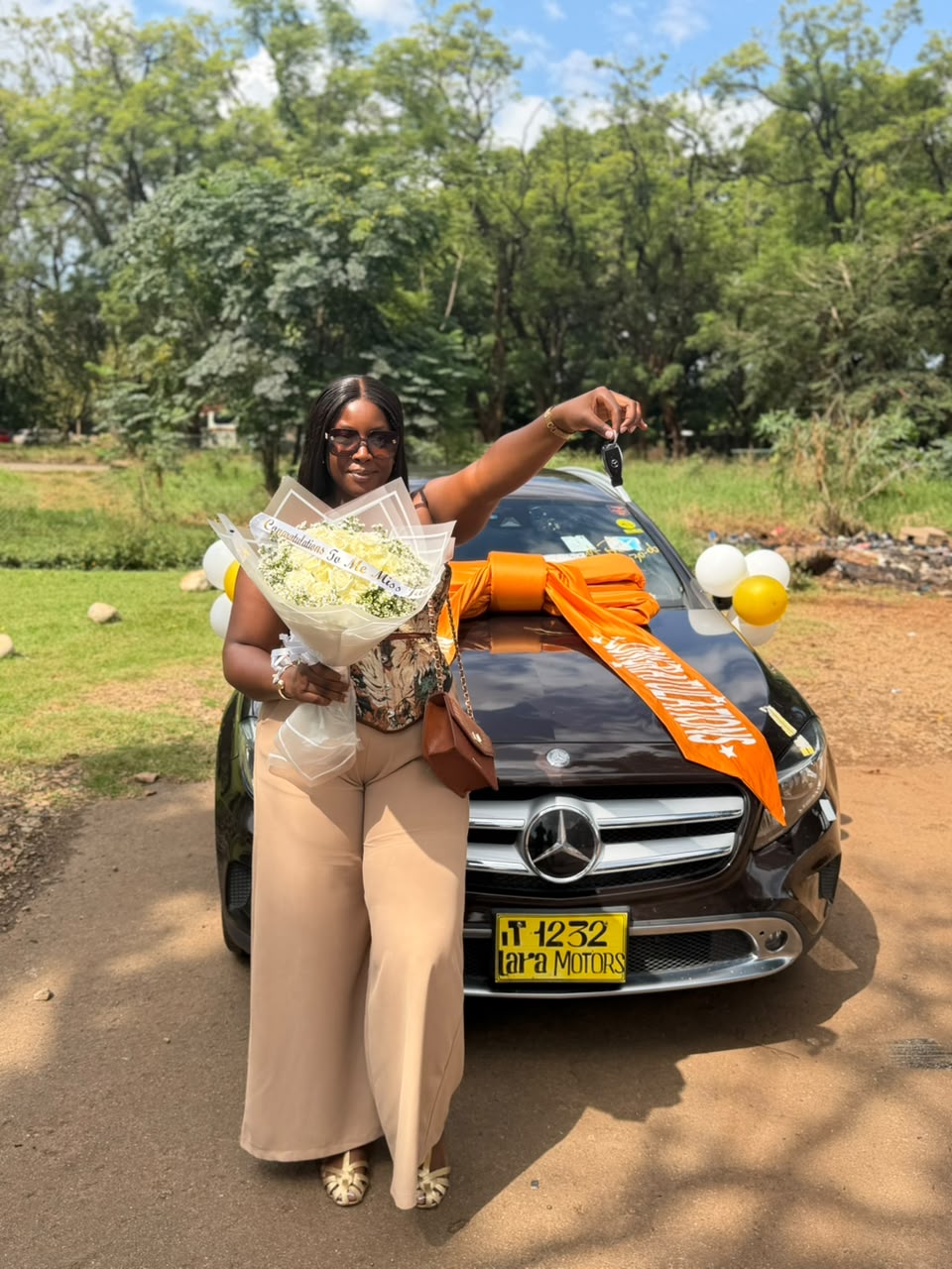 Customer holding flowers and keys beside a brown Mercedes-Benz GLA with orange Lara Motors ribbon